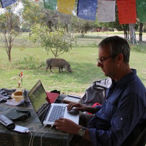 David Bunn on a laptop in South Africa with a Warthog in the background