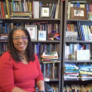 Camille Dungy sitting in front of a bookcase, looking at the camera and smiling