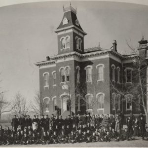 A group of young men and women stand, kneel, sit, or lounge in front Old Main in 1886.
