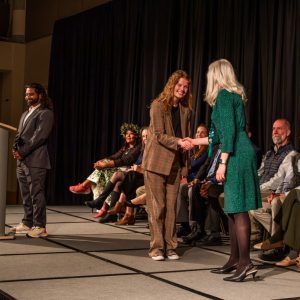 Person in plaid suit shaking hands with person in green dress on stage at Graduate Student Showcase, Nov. 19.