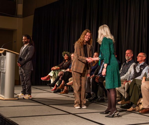 Person in plaid suit shaking hands with person in green dress on stage at Graduate Student Showcase, Nov. 19.