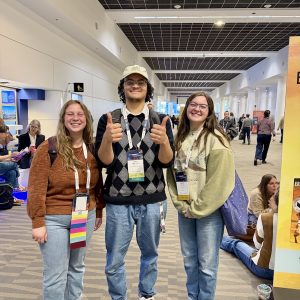 Three students smile and give thumbs up in a convention center hallway with solar panel ceiling visible.