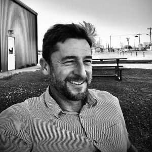 Dan Beachy-Quick sitting outdoors in black and white, wearing a striped shirt, with an industrial background and picnic area.