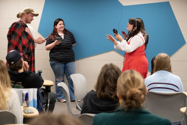 Students perform scenes in front of the classroom, reading lines off their mobile phones