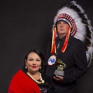A photo of Norma Baker-Flying Horse (Red Berry Woman) on the left in a red shawl and Joe Pekara (Pharaoh 171) standing on the right in a headdress.