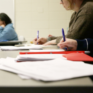 A student wearing a brown sweater sits at a classroom desk, writing with a blue pen on white papers, with a red folder and additional materials spread across the desktop
