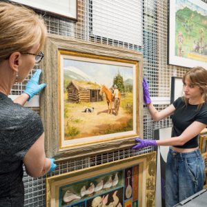 Two women hold a painting as they work to hang it up for display.