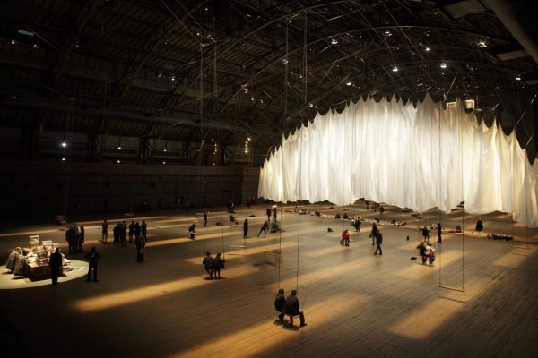 An overhead shot of an art exhibition featuring a large expanse of white cloth and people standing and sitting in front of it.