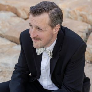 Zach Hutchins in dark suit and patterned bow tie seated outdoors against rocky background, styled in vintage aesthetic.