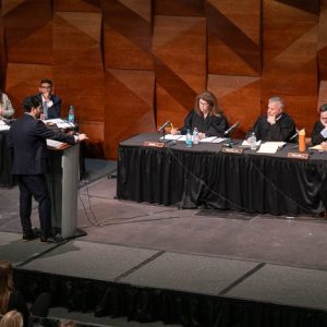A participant speaks at a podium while judges sit at tables during a formal debate or hearing.