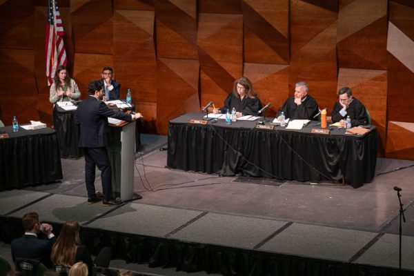 A participant speaks at a podium while judges sit at tables during a formal debate or hearing.