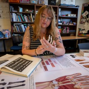 Person studies a skeletal hand model with anatomy books and diagrams spread across a table.