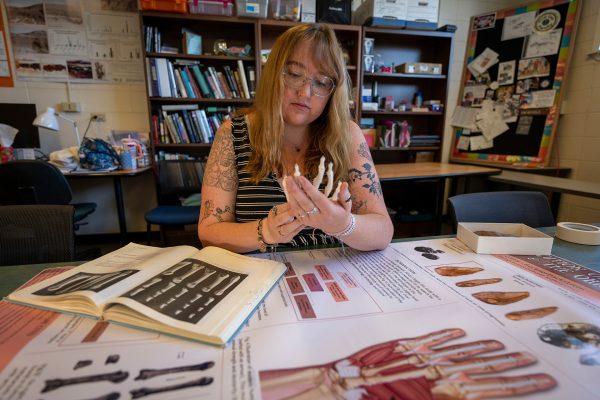Person studies a skeletal hand model with anatomy books and diagrams spread across a table.