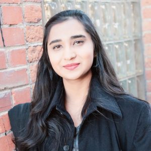 Saira Hafeez poses for a portrait in front of a brick wall and glass-block window.