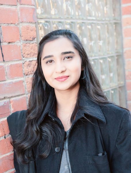 Saira Hafeez poses for a portrait in front of a brick wall and glass-block window.