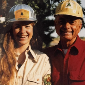 A photo from the 70's depicts a girl with long red hair and her father in a red shirt, both of whom are wearing hard hats that say "Colorado State Forest Service."