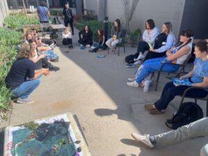 Students sit outside in a circle talking about their class and their cyanotype in Thresholds & Encounters // Meeting the Universe Halfway
