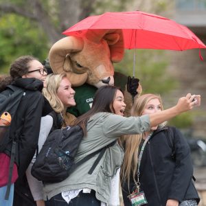Students cheerfully taking a selfie with a mascot under a red umbrella on a rainy day.