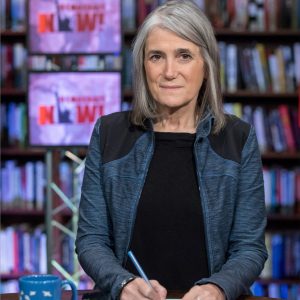 Journalist Amy Goodman sits at a desk holding a pen over a stack of papers. Bookshelves and two screens displaying the Democracy Now! logo are visible behind her.