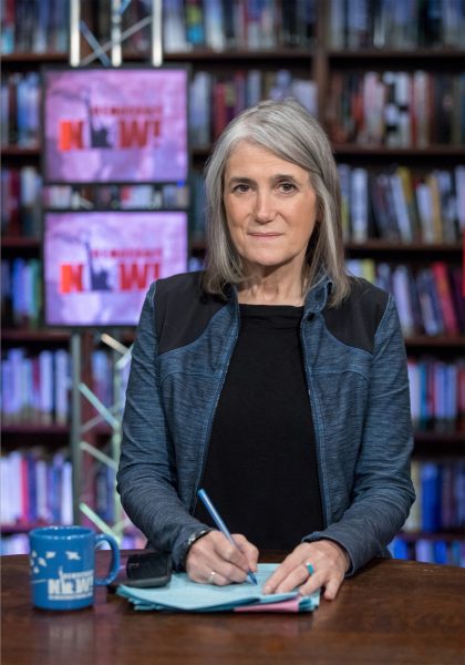 Journalist Amy Goodman sits at a desk holding a pen over a stack of papers. Bookshelves and two screens displaying the Democracy Now! logo are visible behind her.