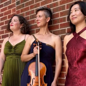 Three women in formal dresses pose against a red brick wall. The woman in the center, wearing a navy blue dress, holds a viola, while the women on either side wear green and burgundy dresses.