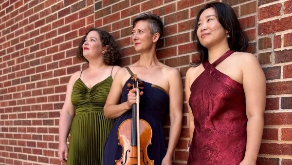 Three women in formal dresses pose against a red brick wall. The woman in the center, wearing a navy blue dress, holds a viola, while the women on either side wear green and burgundy dresses.